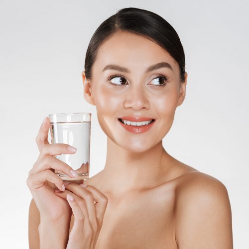 Beauty portrait of young happy woman with hair in bun drinking still water from transparent glass isolated over white background