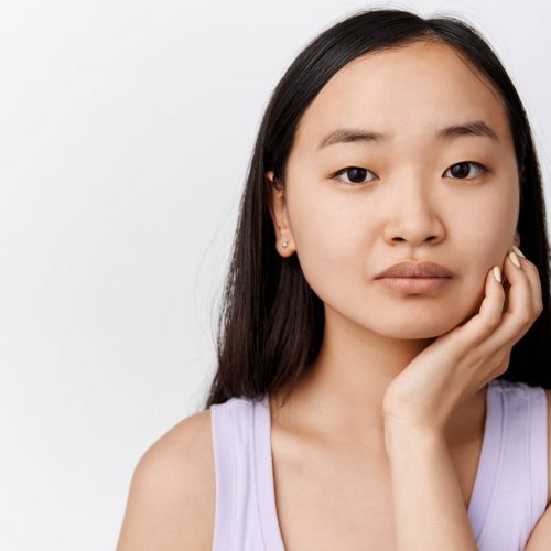 Close up portrait of asian girl touching face and gazing at camera, promo of skin care cosmetic product, cleanser or moisturizer, standing over white background.